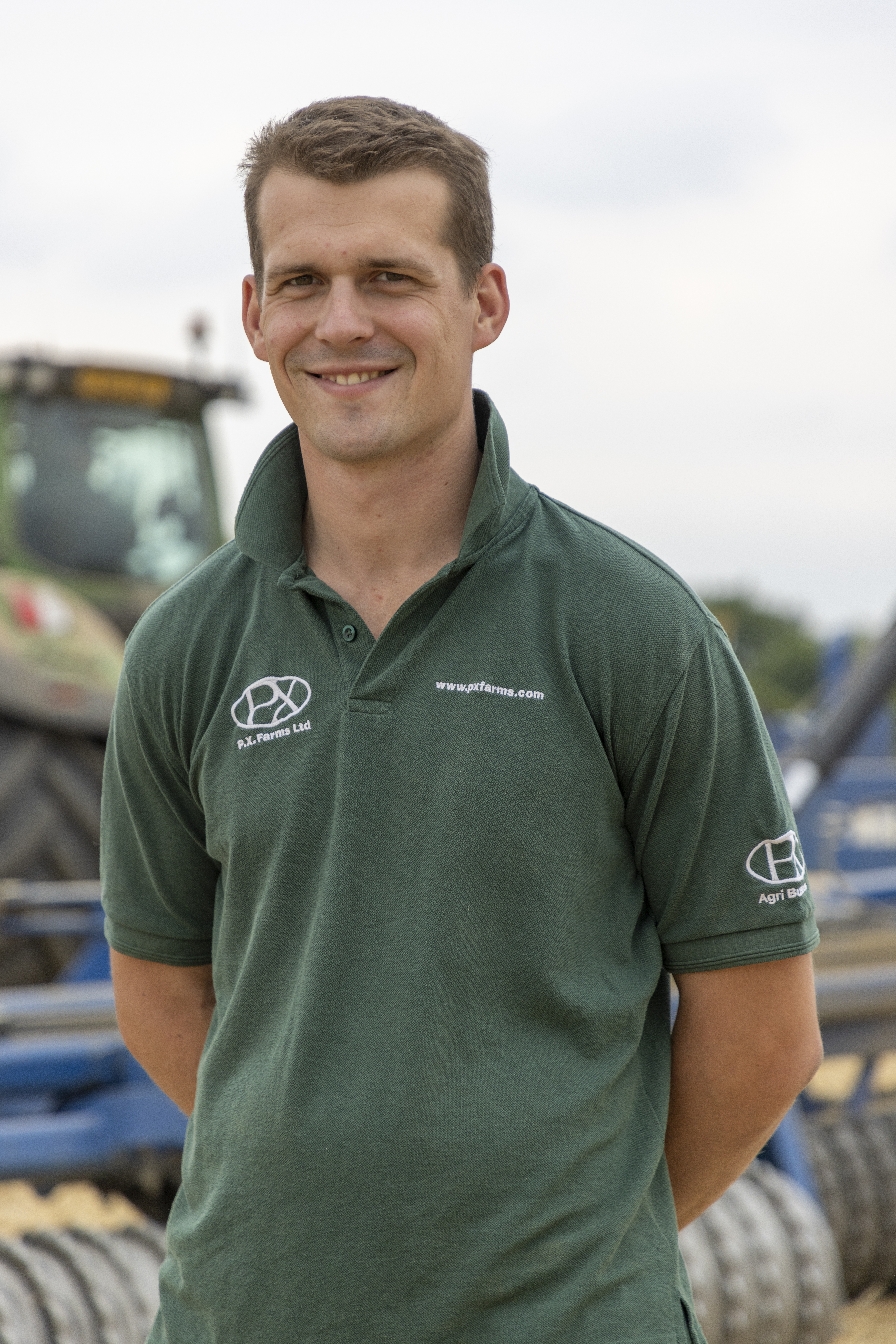 Picture of Tom Eve standing for a photo with his hands behind his back and a tractor in the background