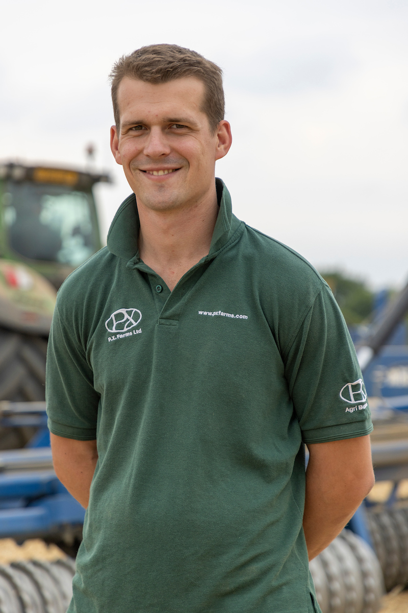 Picture of Tom Eve standing for a photo with his hands behind his back and a tractor in the background
